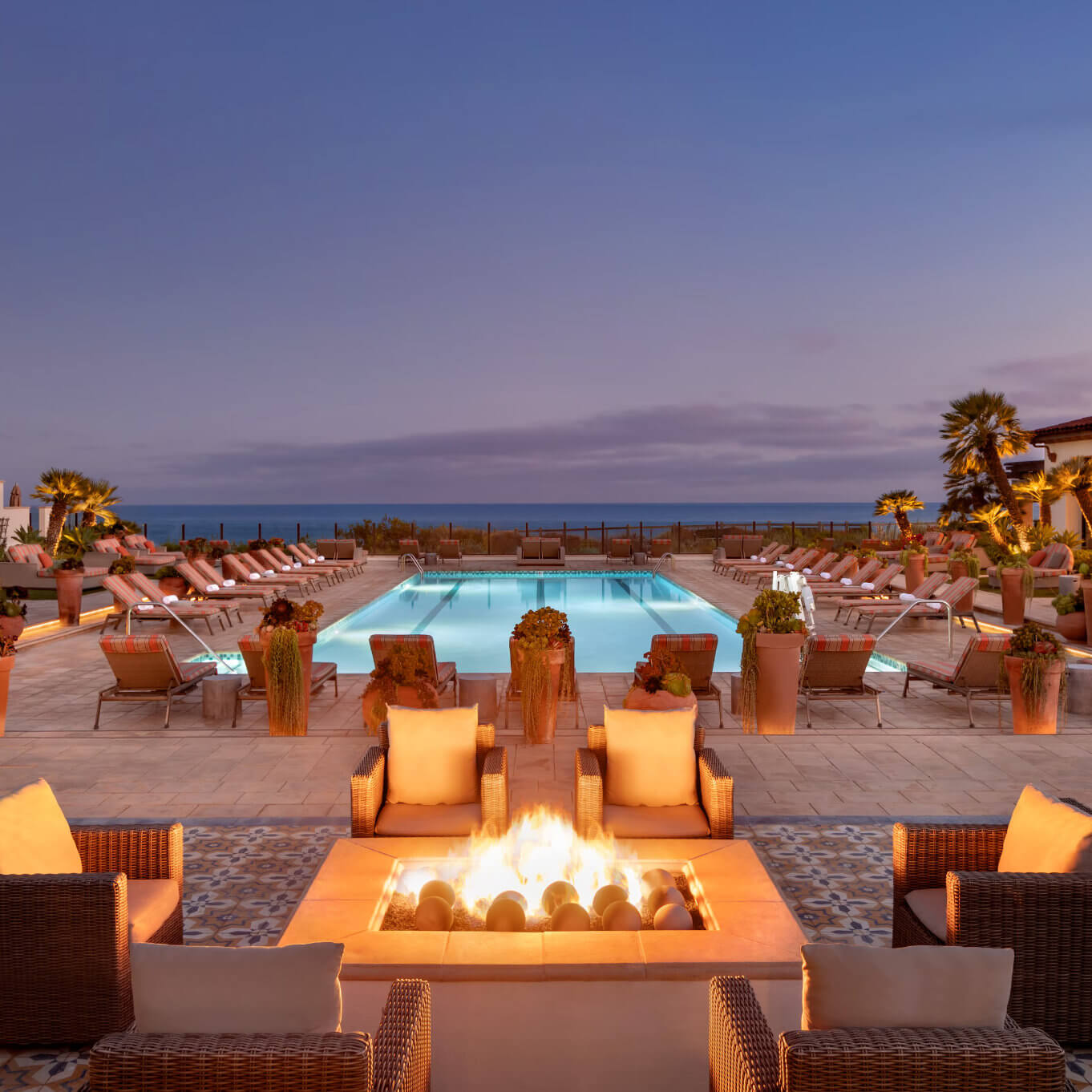 A lit fire pit and lounge chairs beside a pool at Terranea Resort at dusk, with the ocean in the background
